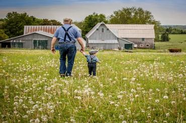 An elderly man holds his grandson's hand as they walk toward a barn
